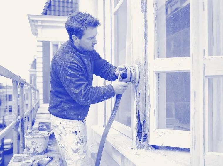 Schilder schuurt op een steiger een houten kozijn, waarbij onder de weggeschuurde verf het houtrot zichtbaar wordt.